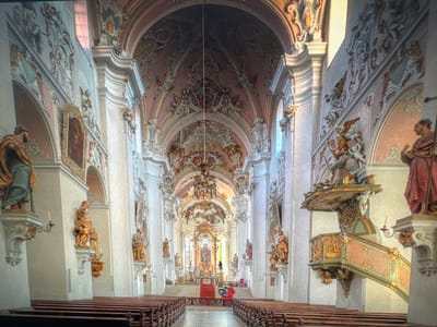 This is an interior view of the St. Vitus Cathedral in Ellwangen, Germany. The image captures the grand nave of the cathedral, looking towards the altar. The architecture is Baroque, characterized by ornate stucco work, gilded details, and vibrant ceiling frescoes. Statues of saints are positioned in niches along the walls. A pulpit, also elaborately decorated, is visible on the right side of the nave. Rows of wooden pews fill the foreground and middle ground. The altar area at the far end is illuminated, drawing the viewer's eye. The scene is devoid of people, creating a sense of stillness and grandeur.