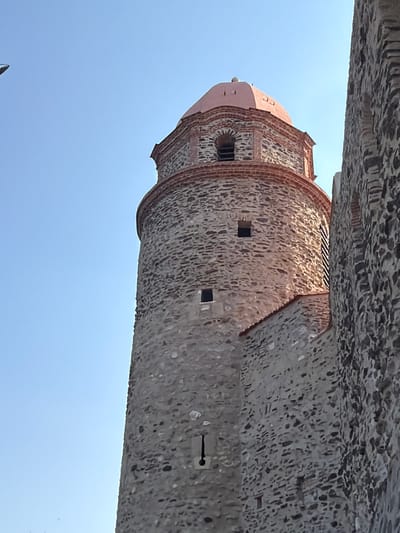 The image shows a close-up, rotated view of a stone tower with a pink dome, possibly a church or part of a historical fortification, against a clear blue sky. The tower is constructed from rough, uneven stones interspersed with reddish brickwork, particularly around the arched openings and the decorative top. Small square windows are visible on the tower's surface. The dome is smooth and rounded, with visible ribs or segments.

Above the tower, a textured stone structure, possibly an overhang or battlement, is visible, composed of irregular grey stones. The composition is tightly framed, with the sky occupying a significant portion of the lower half of the image. The lighting suggests it is daytime, likely late morning or afternoon, given the brightness and clear sky, implying fair weather.

While no people are clearly identifiable, the image's perspective, looking upwards towards the architecture, could imply a viewer standing below. The setting appears to be an outdoor, possibly historical, location, with the architectural style hinting at a European or Mediterranean context. The details of the stonework suggest age and a robust construction. No text is visible in the image. The provided location context of Collioure, France, aligns with the architectural style and possibility of a coastal or historical town setting.