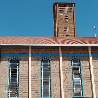 The image displays the exterior of a building, identified as a church, situated in Machakos Central ward, Kenya, against a clear blue sky. The structure features walls constructed from light brown, coursed brickwork. A red-orange corrugated metal roof covers the main edifice, with a white gutter system visible along its eaves. The visible facade includes three tall, narrow, pointed-arch windows. Each window contains multiple horizontal louvered panes set within white frames, outlined by blue trim and minor multi-colored decorative elements. A horizontal blue stripe runs across the brick wall above these windows, and vertical cream-colored architectural elements separate the windows and frame the corners of the building. To the right, a prominent, rectangular bell tower, constructed from matching brick, rises above the roofline. This tower exhibits several rectangular openings with a grid pattern. A small white Christian cross is affixed near the upper right edge of the bell tower. No individuals, actions, or interactions are present in the scene. A "TECNO" watermark is visible in the lower left corner of the image.