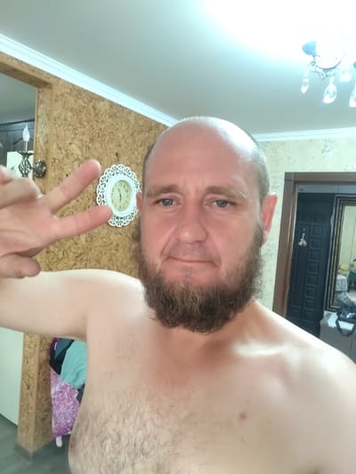 A man with a bald head and a beard is taking a selfie. He is shirtless and holding up two fingers in a peace sign. In the background, there is a clock on the wall, a chandelier on the ceiling, and a doorway. The image appears to be taken indoors, possibly in a residential setting. There is no information about the location in Petropavl City Administration, Kazakhstan.