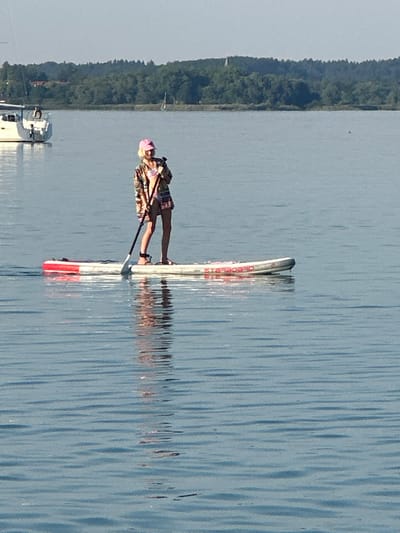 This media file captures a person stand-up paddleboarding on a calm body of water. The individual is wearing a camouflage-patterned top, plaid shorts, and a pink headband, with a paddle in hand. The paddleboard itself is white with red accents. The water reflects the paddleboard and the person, creating a mirrored image. In the background, a tree-lined shore is visible, with a few boats docked near the edge of the water, one of which appears to be a sailboat. The scene suggests a peaceful outdoor activity, likely taking place during daylight hours given the bright lighting and clear reflections. The overall impression is one of a leisurely day spent on the water. There are no discernible emotions evident from the subject's posture or attire. Some text is visible on the paddleboard, appearing to be red lettering. Based on the location context provided, this scene is likely Gstadt am Chiemsee, Germany.