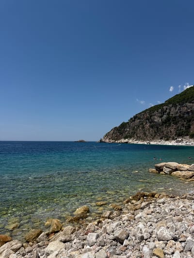 The image depicts a view of the coastline at Perazića Do, Montenegro. The scene is dominated by a clear, blue sky and the turquoise waters of the Adriatic Sea. The foreground is a rocky beach with numerous grey and beige stones of various sizes. Some larger rocks are present near the water's edge.
The water is remarkably clear, allowing visibility of the submerged rocky seabed. In the middle ground, several people are swimming and wading in the sea. Further out, the water transitions to a deeper, darker blue.
A prominent feature of the background is a steep, green-covered mountain slope that curves along the coastline. The mountain is textured with rock faces and patches of vegetation. A small, rocky islet is visible in the distance, further out in the sea, with a line of buoys or markers extending from the shore towards it. In the far distance, the coastline appears to extend further, with a small beach area populated by numerous sunbathers. A few wispy clouds are present in the upper right corner of the sky.