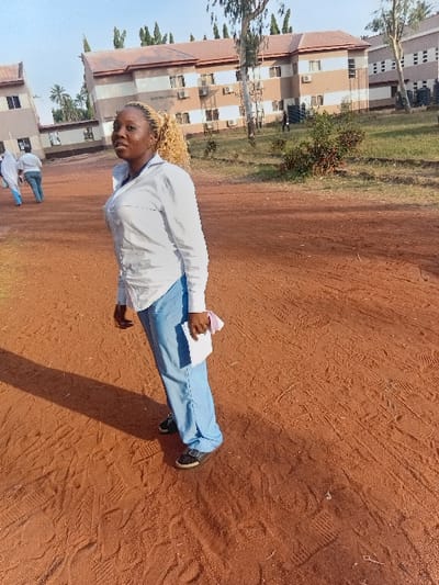 The image captures an outdoor scene, likely on a campus or institutional ground, featuring a young person as the main subject. The perspective is from a slightly elevated angle, looking across a wide, unpaved area towards a series of buildings.
The central figure is a young individual, appearing to be female, standing towards the right side of the frame. She has light-colored, curly hair, possibly blonde or light brown, and is looking directly at the camera with a slight smile. She is wearing a white long-sleeved top, which appears to be a uniform or scrub-like attire, and light blue trousers. Her shoes are dark-colored. In her left hand, she holds a stack of white papers or a small notebook. Her right hand is near her chest, possibly holding a small pink object or gesturing. A distinct shadow extends from her body onto the ground behind and to her right.
The ground covering the foreground and mid-ground is a reddish-brown earth, characteristic of dirt or clay. Numerous footprints and tire tracks are visible on the surface, indicating frequent passage.
In the background, several multi-story buildings are visible. These buildings share a consistent architectural style, featuring light brown or beige facades with multiple rectangular windows and dark reddish-brown sloped roofs. Many of the windows have white-colored air conditioning units mounted beneath them. Tall, slender trees with green foliage are scattered between the buildings and on the periphery of the open area. Some smaller bushes are also visible near the building structures.
Further in the background, towards the left, two other individuals can be seen. They also appear to be wearing similar white tops and light blue pants, suggesting they are part of the same group or organization as the main subject. They are walking or standing further away from the camera.
The scene is brightly lit by natural sunlight, indicating it is daytime, likely during the mid-morning or afternoon, given the length and direction of the shadows. The sky is not visible in the frame. No discernible text is visible in the image.