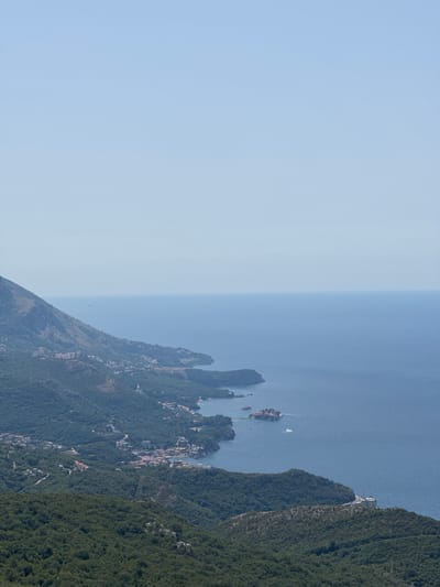 This is a high-angle, wide shot of a coastal landscape in Brajici, Montenegro. The scene is dominated by a vast expanse of deep blue water meeting a clear, pale blue sky. In the mid-ground, a lush, green mountainside slopes down towards the coast, dotted with buildings that form a coastal town. A serpentine road winds its way along the mountainside, and a few structures are visible nestled within the greenery. Further out at sea, a small island, possibly Sveti Stefan, is visible, connected to the mainland by a narrow causeway, with a white yacht sailing nearby. The foreground is filled with the dense, dark green foliage of a forested mountain. The overall impression is one of a serene, sunlit day with a panoramic view of the Adriatic coastline.