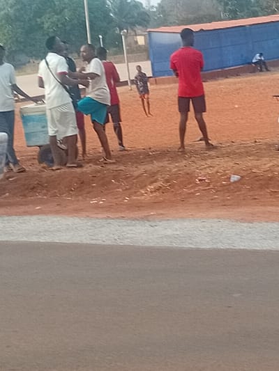 The image depicts an outdoor scene in Ayangba, Nigeria, featuring an unpaved reddish-brown dirt area next to a paved gray road. Seven individuals are present. In the left-foreground, a person in a white t-shirt and white shorts, carrying a dark shoulder bag, stands adjacent to a light-colored cooler on wheels. Immediately to their right, a second individual in a light-colored t-shirt and blue shorts is captured in mid-motion with their left leg raised and bent, facing the first person. To the right of these two, a third person in a red t-shirt and dark shorts stands with their back to the viewer. Further back, a fourth person in a red t-shirt and dark shorts is partially visible. A prominent blue rectangular building with a red top trim stands in the mid-background to the right. A fifth individual in a patterned dark shirt and shorts stands on the dirt in front of the blue building. In the far background, a sixth person in a white shirt and dark pants is seated near the base of the blue building. Two slender light poles are visible in the background to the left of the building. Distant green foliage frames the upper background. The dirt ground contains scattered small white debris. The lower portion of the image shows a gray paved road with a distinct lighter gray edge marking the transition to the dirt area.