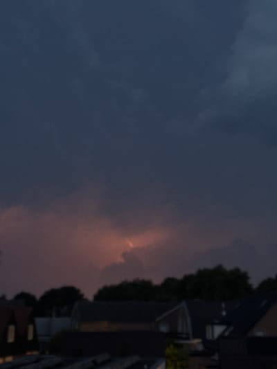 The image captures a twilight scene in Putten, Netherlands, looking towards the horizon. The sky is dominated by dark, heavy clouds, with a sliver of dramatic orange and yellow light breaking through in the upper-middle section, suggesting a sunset or perhaps a distant light source. Below the sky, the silhouettes of trees and residential rooftops are visible. The architecture suggests a suburban or semi-urban setting, with peaked roofs and dark facades. The overall mood is somber and atmospheric, hinting at an approaching storm or the end of a day.