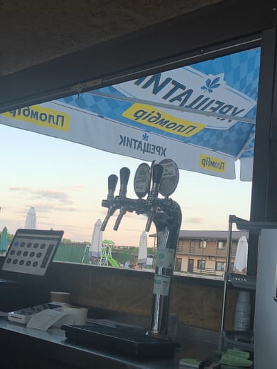 The image depicts a bar counter at an outdoor establishment, likely during twilight. The focal point is a beer tap with multiple spigots, positioned in the foreground. To the left of the taps is a point-of-sale system with a screen and keypad, alongside a receipt printer and other small payment devices. In the background, a large blue and white checkered umbrella provides shade, with a logo featuring a leaf motif and text in what appears to be Cyrillic script. Beyond the bar, the scene opens to an outdoor area with white umbrellas, a fence, trees, and buildings with wooden facades. The sky exhibits soft, pastel hues, suggesting a sunset. The overall impression is a service area at a recreational venue, possibly a restaurant or cafe, in the city of Shkurupiivka, Ukraine.
