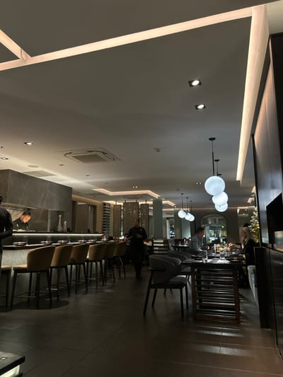 A dimly lit restaurant with a long, dark wooden table stretches into the distance. Modern spherical light fixtures hang overhead, illuminating the bar area where a few patrons sit. Waitstaff are visible at the ends of the table. Square-backed chairs line the table, some upholstered in dark fabric and others in a tan material. The walls are light-colored with recessed lighting and vents. The overall ambiance is sophisticated and modern.