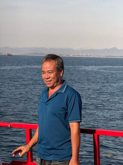 A medium shot captures an East Asian man in his 50s standing on a boat deck, facing right, with the ocean and a distant coastline in the background. He has short, graying hair, fair skin, and is smiling slightly, with his mouth open. He wears a dark blue collared shirt with white trim on the collar and sleeves, and dark gray patterned shorts. His left arm is bent at the elbow, and his left hand is extended forward, pointing. A bright red railing runs horizontally in the foreground, with a vertical support post visible on the left side of the frame. The water is a deep blue, with visible ripples. In the far distance, a hazy coastline with mountains is visible, along with some structures and a small boat on the left. The sky is a pale, hazy blue. The location is identified as An Hải, Vietnam.