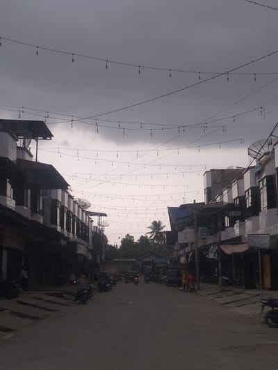 The image depicts a street scene in Keudegeudong, Indonesia, under a heavy, overcast sky. The street is lined with multi-story buildings, some appearing to be shops or residences, with awnings and signage visible, including "TACO". String lights are suspended across the street, illuminated by the dim ambient light. A few motorcycles are parked along the roadside and a couple are in motion on the asphalt road. Several people are present, some standing near the buildings and others on motorcycles. The environment suggests a marketplace or a commercial area, possibly experiencing a decline in activity due to the weather or time of day.