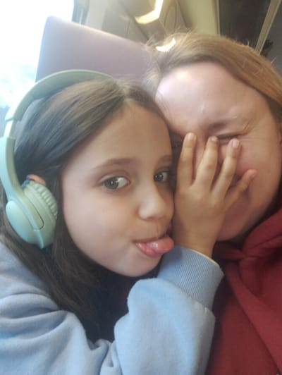 The image displays two individuals, an adult female and a female child, situated within a transportation vehicle, likely a train, in Avignon, France. The adult, positioned on the right side of the frame, has reddish-blonde hair and wears a red hooded garment. Her face is partially obscured by the child's left hand, which covers her left eye and the bridge of her nose. Her visible right eye is squinted, consistent with a smile or laugh. The child, on the left, has dark hair and is wearing light blue over-ear headphones. She is dressed in a light blue sweatshirt. Her tongue is extended outwards, and her left hand is pressed against the adult's face. The background reveals portions of a purple upholstered seat and a window admitting bright external light, along with overhead interior light fixtures typical of a train carriage.