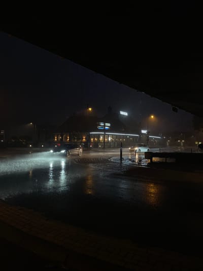 A low-angle, wide shot captures a dark, wet street at night in Staphorst, Netherlands. The foreground shows a wet, brick sidewalk and a street covered in puddles, reflecting the surrounding lights. In the mid-ground, a silver car drives through a large puddle, creating a significant splash of water illuminated by its headlights. Across the street, a building with a prominent "WANDERS" sign is visible, its windows lit and reflecting on the wet surface. Streetlights cast a yellowish glow on the scene. The overall atmosphere is dark and rainy, with the water on the ground and the spray from the car suggesting recent or ongoing rainfall.
