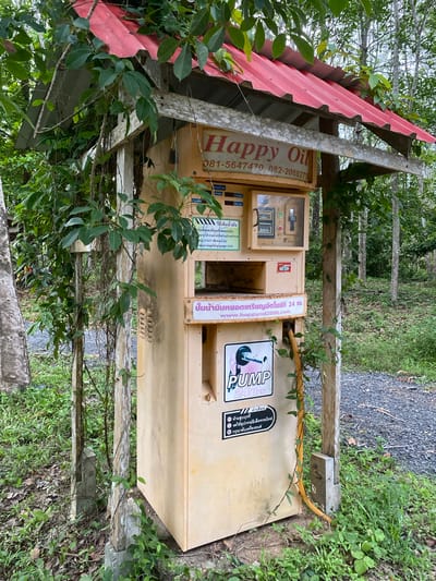 A weathered, pale yellow, automated gasoline pump structure stands outdoors under a red corrugated metal roof supported by wooden beams and posts, some of which are entwined with vines and foliage. The pump has signage on its front, including "Happy Oil" in red lettering and Thai script. Below this, a digital display and slot are visible within a glass-enclosed compartment. Another section of the pump features a sticker with a cartoon hand holding a nozzle and the word "PUMP 24Hrs." A yellow hose hangs from the pump. The structure is situated on a gravel path surrounded by lush green grass and trees, suggesting a rural or semi-rural setting. The overall impression is one of a functional, albeit aged, self-service gasoline station operating in a natural environment. The image was taken in the city of Ko Siboya, Thailand.