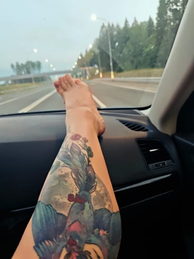 A person's leg with a large tattoo of roosters and a moon is resting on the dashboard of a car. The car is driving on a highway near DubrovKI, RUSSIA, as indicated by the trees and streetlights in the background. The sky is overcast.