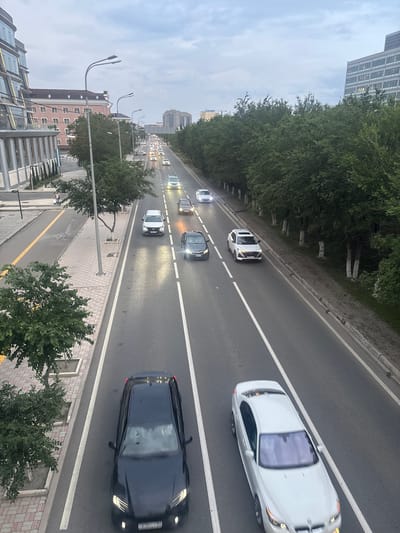 This image depicts a street scene in Atyrau, Kazakhstan. The photo is taken from an elevated perspective, looking down at a multi-lane road with traffic. Several cars are visible, including a white SUV in the foreground and a black sedan behind it. Further down the road, a line of cars, mostly white, are moving in the same direction. The road is lined with trees and a sidewalk. In the background, buildings and more trees can be seen. The sky is overcast, suggesting it may be daytime with cloudy weather. The lighting indicates it is likely late afternoon or early evening, as car headlights are illuminated. There are no people clearly visible in the image. The scene is of everyday urban traffic.