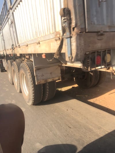 A partial view of a semi-trailer truck is shown from the side. Multiple tires are visible on the truck's axles, along with parts of the trailer's frame and body. The truck appears to be on a paved surface, possibly a road or parking lot. The trailer body is constructed from corrugated material, likely metal. The truck appears to be older, judging by the rust.