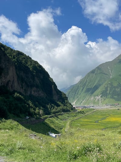 This image depicts a mountainous landscape in Kanobi, Georgia. The foreground is dominated by a grassy slope dotted with wildflowers. A dirt path winds its way down the slope. In the midground, a paved road runs along the base of a steep, tree-covered mountain on the left. A white vehicle is parked on the side of the road. Across the valley, on the right, is another large mountain, green and sparsely covered with trees, with patches of snow visible on its higher reaches. In the distance, a small village or town is nestled at the foot of the mountains. The sky is a vibrant blue with large, puffy white clouds.