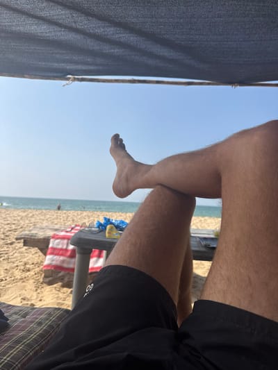 A low-angle perspective of a person sitting near a beach. Hairy legs are crossed in the foreground with one foot dangling in the air. Black shorts are visible on the right. A dark fabric shade structure is on the left. In the background, the beach is visible, with sand, the ocean, a striped towel, and a table visible. The sky is clear and bright.