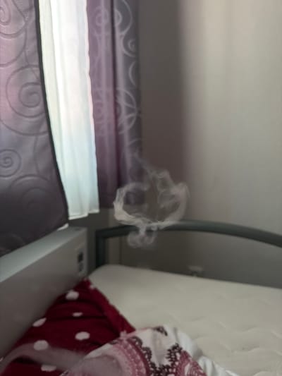 The image depicts a close-up view of a bed with layered bedding. The bedding appears to be in shades of purple and white, with patterned textures. A wisp of white smoke is visible in the upper center of the frame, swirling in the air. To the right of the bed, a white object, possibly a bedside table or piece of furniture, is partially visible, along with some fabric with a white polka dot pattern and decorative trim. The overall lighting suggests an indoor setting, potentially in a bedroom. The activity taking place is not explicitly clear due to the focus on the bedding and the presence of smoke, but it could imply someone has recently smoked or used an item that produced smoke in the room. No specific time of day or weather conditions can be inferred. There is no visible text in the image. The setting appears to be a domestic environment.