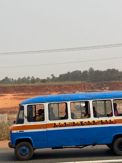 The image displays a blue, white, and orange passenger bus traveling on a paved road in Orifite, Nigeria, under a hazy, light-colored sky. The word "MERCIFUL" is prominently displayed in orange and red lettering on the bus's side. A male driver, wearing a yellow shirt and a dark cap, is visible in profile in the driver's seat. Multiple passengers are seen through the windows, primarily as dark silhouettes, with one individual in red/pink attire partially visible towards the rear. In the mid-ground, a large excavation site with reddish-brown earth is evident, containing several light-colored vehicles or machinery and scattered concrete blocks. Beyond this site, a dense line of tropical trees, including palms, forms the horizon. Three horizontal power lines traverse the upper portion of the sky.