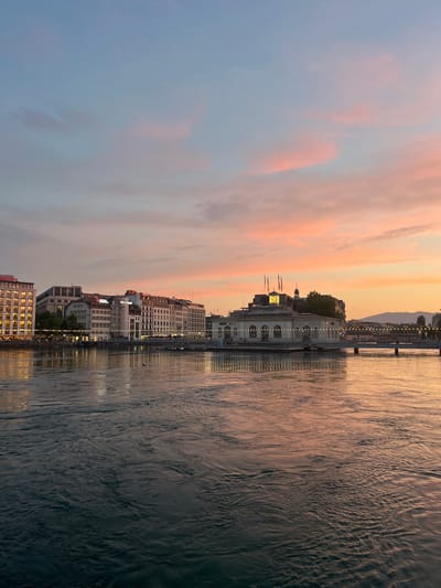 This image captures a waterfront view of Geneva, Switzerland, during what appears to be a sunset. The sky is painted with soft hues of pink, orange, and blue, with scattered clouds. In the foreground, a body of water, likely Lake Geneva, reflects the warm colors of the sky and the lights from the city.

Dominating the mid-ground is a row of buildings lining the waterfront. The architecture suggests a mix of older and more modern structures. One prominent building in the center, with a clock face and flags, appears to be a public or commercial building. The buildings are illuminated with warm, yellow lights, creating a welcoming atmosphere. String lights are also visible, adding to the festive ambiance along the waterfront promenade.

A bridge spans the water in the background, connecting the city. Distant hills or mountains can be seen on the horizon, silhouetted against the colorful sky. There are a few individuals visible walking along the waterfront, suggesting it is a public space. The overall scene conveys a sense of tranquility and urban beauty at dusk.