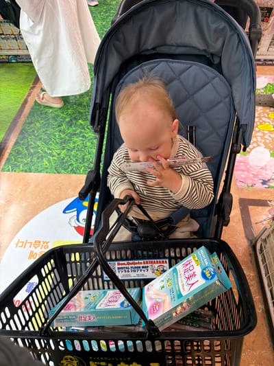 A Caucasian toddler, wearing a striped long-sleeved shirt and tan pants, sits in a blue stroller. The toddler is holding a small rectangular object to their mouth. The stroller is placed within a black shopping basket containing several boxes of "Cool Refresh Sheet" products. The background shows a colorful floor with a cartoon character and floral imagery, suggestive of a retail environment, possibly in Chiyoda, Japan, given the presence of Japanese text on the product packaging. A person wearing a white skirt and sandals is partially visible to the left.