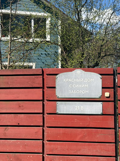 A close-up shows a red wooden fence with two gray plaques. The plaque on the left reads "Красный дом с синим забором", which translates to "Red house with a blue fence". The plaque on the right reads "21 B". A glimpse of a blue wooden house is visible behind the fence and a partially visible tree, suggests the season is spring. The image was taken in Янино-2, Russia.