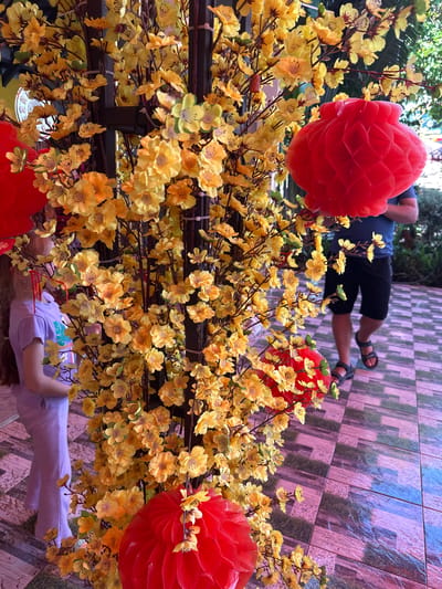 A young girl with long blonde hair wearing a light purple t-shirt and pants stands to the left of a decorative tree. The tree is adorned with numerous small yellow flowers and three red, spherical paper lanterns. The girl is facing away from the camera and towards the tree. In the background, a man wearing a blue t-shirt and black shorts is walking away from the camera and to the right. He is wearing dark sandals. The ground is tiled with a pattern of brown and beige squares, with some areas appearing wet and reflective. The scene appears to be outdoors, possibly in a courtyard or patio area, given the tiled ground and the presence of greenery in the background. The overall impression is one of festive outdoor decoration, likely for a holiday or celebration. The location is in Ia Hru, Vietnam.
