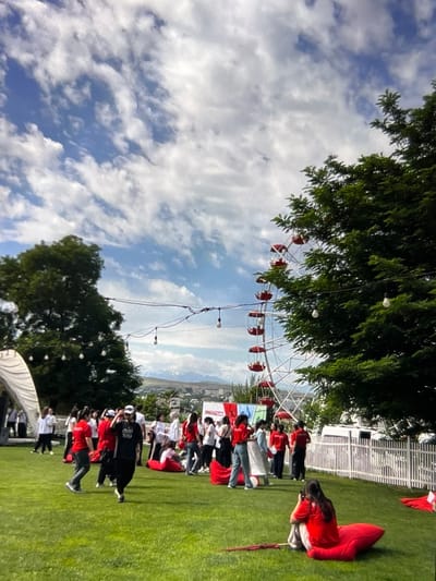 The image depicts an outdoor gathering in a grassy area with a Ferris wheel in the background. Several people are visible, many wearing red and white attire, interacting in various ways: some are standing and talking, others are sitting on large red beanbag chairs. A Ferris wheel with red gondolas is a prominent feature in the mid-ground, partially obscured by a large green tree on the right. String lights are strung across the scene, adding to the festive atmosphere. In the distance, a city skyline and mountains are visible under a sky with scattered clouds. The location is identified as Kadamjay, Kyrgyzstan.
