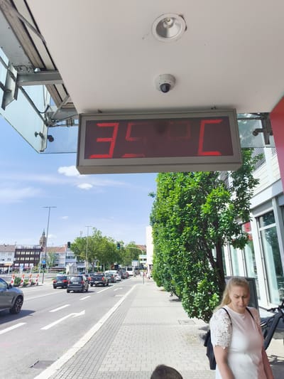 The image shows a street scene in Heilbronn, Germany on a sunny day. A digital thermometer mounted on a white overhang displays a temperature of 35°C. Below the thermometer, a sidewalk runs alongside a street with traffic. A blonde woman in a white lace-sleeved dress is walking on the sidewalk, looking down. A black bicycle is parked near her. On the street, cars are driving in the same direction. In the background, there are buildings, trees, and traffic lights. A church spire is visible in the distance. The sky is blue with some white clouds.