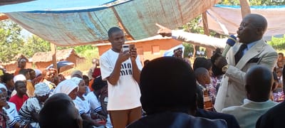 The image shows a gathering of people in what appears to be a rural area of Kalemie, Democratic Republic of the Congo. The setting is outdoors, possibly under a makeshift canopy of blue tarpaulin. Traditional houses with thatched roofs are visible in the background.
A man in a light-colored suit, holding a microphone, is speaking or preaching while gesturing with his right hand. He is wearing a watch on his left wrist. A younger man is standing nearby, holding up a smartphone, seemingly taking pictures or recording video.
Several individuals are seated, mostly women wearing traditional clothing and head coverings. The composition suggests a large crowd present. A few children are among the crowd.