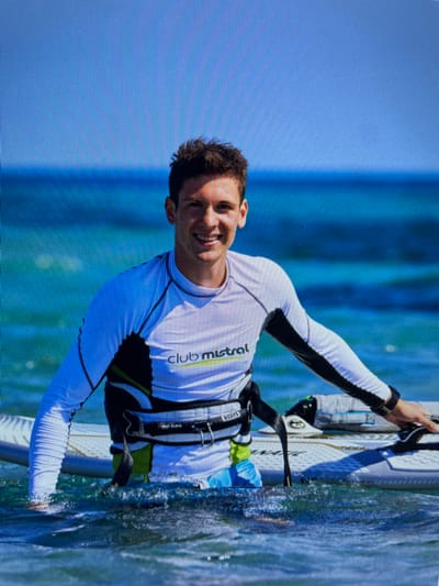 A smiling man is windsurfing in Bacong, Philippines. He's wearing a white long-sleeved shirt with black accents and a black harness. He is holding onto the windsurfing board, which is partially submerged in the water. The water is turquoise, and the sky is light blue. The Club Mistral logo is visible on his shirt.