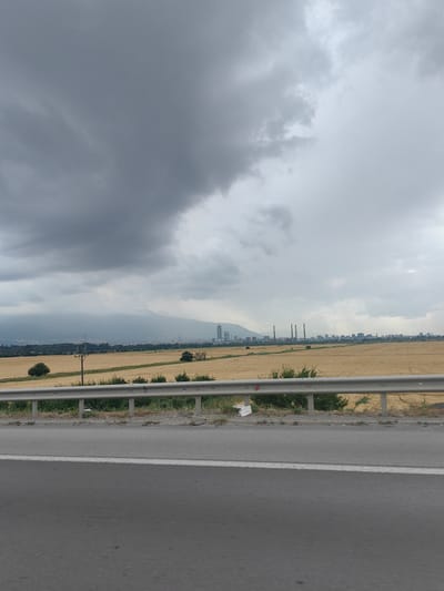 The image captures a wide, horizontal view of a landscape under a heavily overcast sky. The foreground is dominated by a grey asphalt road with a white line marking its edge. Immediately beyond the road is a metallic safety barrier, behind which lies a golden field of dry crops. Scattered green bushes and a few small trees are visible within and at the edge of the field.

In the mid-ground, a power pole with wires stands tall in the field. Further in the distance, a hazy mountain range forms the backdrop. A city skyline is visible along the horizon, characterized by several tall buildings and prominent industrial chimneys, suggesting a power plant or factory. The sky is filled with dark, heavy clouds, indicating a gloomy or stormy weather condition. The overall mood is somber and industrial, with the natural landscape juxtaposed against urban and industrial elements. There is no visible text in the image. The time of day appears to be late afternoon or early evening, given the dim lighting and cloud cover. The location context suggests Kazichene, Bulgaria.