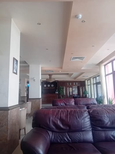 The image shows the reception and restaurant area of a hotel. In the foreground, there are dark brown leather couches. Behind the couches, there is a reception desk with "RECEPTION" written on it, and a "RESTAURANT" sign. There are chandeliers and clocks visible above the reception desk. The room has large windows with light curtains. The environment appears to be indoors and is well-lit. There are no people visible in the image.