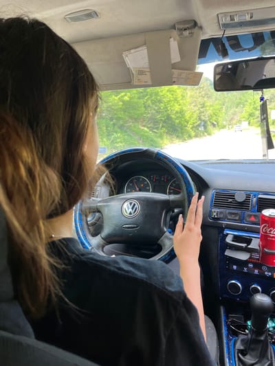 The image depicts the interior of a car, viewed from the passenger seat, with a person's head and shoulders visible from the back. The driver, a young female with long brown hair, is behind the steering wheel, her left hand resting on it and her right hand extended slightly forward. The car's dashboard is visible, featuring a Volkswagen logo on the steering wheel, a speedometer, and various controls. A can of Coca-Cola is placed in a cup holder on the right side of the dashboard. Outside the car, a road is visible, lined with green trees, suggesting a rural or suburban setting. The location is identified as Vlahovo, Bulgaria.