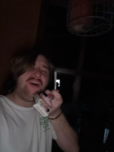 A man with long hair and a beard holds a cigarette to his lips, a pack of Marlboro cigarettes against his face, and a lighter in his hand. He is wearing a white t-shirt and a bracelet on his wrist. A decorative wheel is visible in the background. The image was taken at night. The location is Cañada de Madero, Mexico.