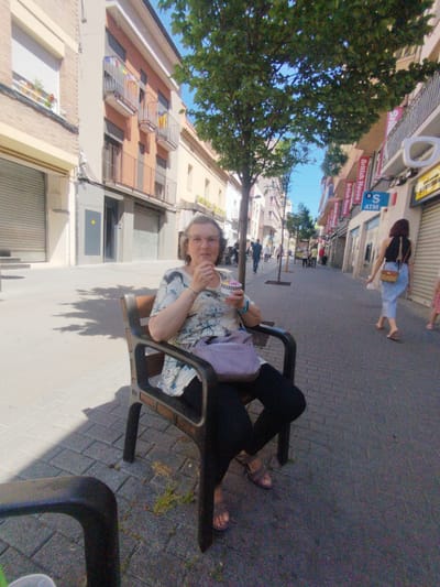 In Rubí, Spain, an elderly woman with glasses sits on a metal and wood chair along a pedestrian walkway. She's wearing a floral print top and dark pants, and is eating from a small container, possibly ice cream. A purple purse rests on the chair beside her. Behind her, the street is lined with buildings of various heights and colors. Storefronts are visible on the right, including one with a "British House" sign and an ATM. People are walking in the background. A tree provides shade over the woman and the walkway. The ground is paved with grey bricks. The weather appears to be sunny.