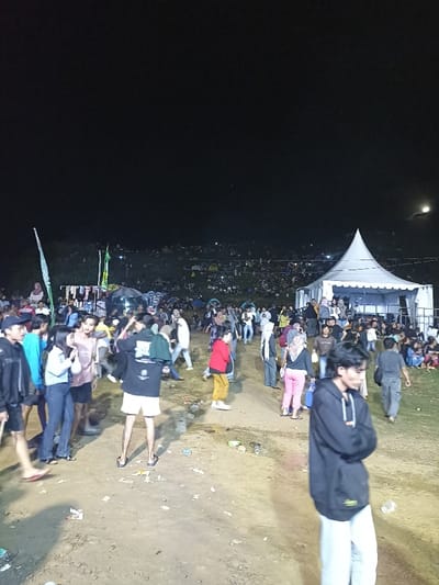 This nighttime image depicts a lively outdoor gathering, likely a festival, concert, or camping event, in Kecamatan Pujut, Indonesia. The scene is illuminated by artificial lights, casting shadows and highlighting the numerous people present.
In the foreground and midground, a large crowd of people, mostly young adults, are seen standing, walking, and socializing on a dirt-covered ground. Many are dressed in casual attire, including hoodies, t-shirts, and jeans. Some individuals are facing the camera, while others are seen from the back or side, moving through the space. The ground shows signs of heavy foot traffic and some scattered litter.
To the right, a large, white, pointed event tent (similar to a pagoda tent) stands prominently, appearing to be a central structure for the event, possibly housing a stage or vendor area. On the left side of the midground, there's a smaller stall or vendor setup with items displayed, and two green flags are visible.
The background reveals a hillside covered with many small lights, indicating a vast expanse of people or numerous camping tents spread across the terrain. The overall impression is a large-scale outdoor overnight event. The sky is dark, characteristic of nighttime, with a faint moon visible in the upper right quadrant, suggesting clear weather. No discernable text is visible on signs or clothing within the image.