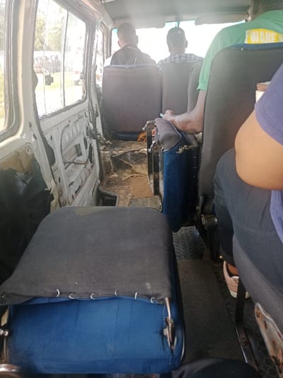 The image displays the interior of a utilitarian minibus taxi, a common form of public transport in Soweto, South Africa. The view is from a rear passenger seat, looking forward into the cabin. Four individuals are present: a person partially visible in the immediate foreground right wearing a dark top; a passenger in a green polo shirt with yellow text on the back, seated mid-ground right, whose right hand grips the seat in front; and two additional passengers seated further forward, their backs to the camera, one in a dark shirt and cap, the other in a plaid-patterned shirt. The vehicle's interior exhibits considerable wear; the left-side paneling is absent, revealing the white metal frame and internal structures. The central aisle floor is scuffed, exposing bare metal in areas and featuring a black rubber mat with circular indentations on the right. The immediate foreground seat has a dark blue base cushion and a dark grey fabric cover secured by metal rings. Natural light illuminates the cabin from the windows, through which stacked tires are visible in an outdoor setting.