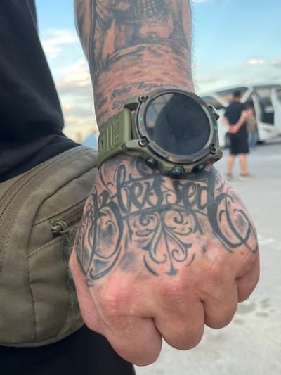 A close-up, low-angle shot focuses on the left hand and wrist of a person, featuring a large tattoo on the back of the hand that reads "Blessed" in ornate script. The person is wearing a green tactical watch on their wrist, with the words "MODE" and "HOME" visible on the bezel. A portion of a dark shirt and a khaki tactical bag are visible on the left side of the frame. In the background, out of focus, two individuals are standing near the open doors of a white vehicle, possibly a van or bus, under a cloudy sky. The setting appears to be an outdoor, possibly urban or semi-urban environment, given the presence of the vehicle and pavement. The image does not contain any specific visual indicators to pinpoint its location in Palanca, Moldova.