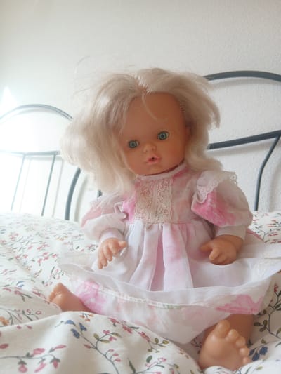 A doll with blonde hair and blue eyes is sitting on a bed with a floral-patterned comforter. The doll is wearing a white and pink dress with lace trim. The bed is made with white sheets and a headboard. The room appears to be a bedroom. The image was taken in Navaluenga, Spain.
