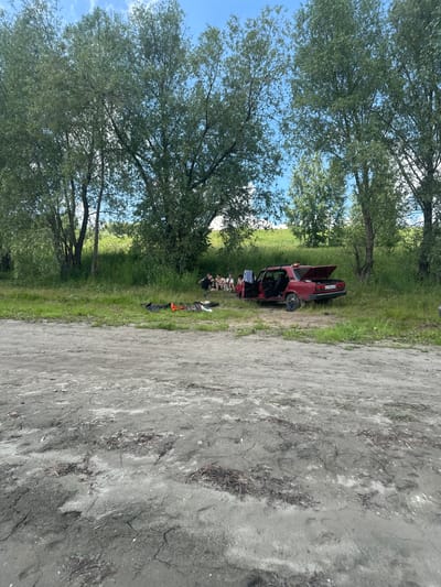 In a grassy area beside a dirt path, a red car with its hood and doors open sits parked. Several people, appearing to be in their late teens or early twenties, are gathered around the car. Some are sitting on the ground, while others stand, seemingly engaged in conversation or taking a break. Various items are scattered around the car, including what looks like striped blankets or towels, a bag, and an orange tool or piece of equipment. The scene is set under a bright, possibly midday sky, with lush green trees providing a natural backdrop. The overall atmosphere suggests a casual outdoor gathering, perhaps a picnic or a stop during a road trip. The license plate of the car is partially visible, displaying Cyrillic characters and numbers, reading "E019BH 777". The location is identified as Pravda, Russia.