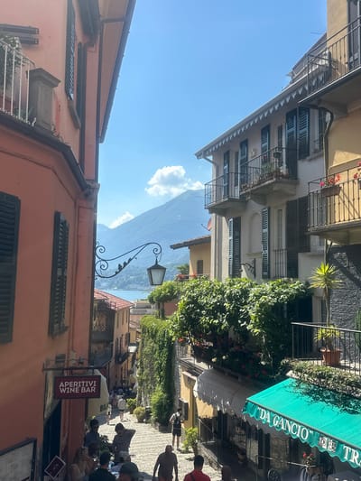 The image depicts a street scene in San Giovanni, Italy, on a sunny day. The orientation of the photo is rotated, but the visual elements suggest an outdoor setting with buildings lining a pedestrian walkway.
The subjects include several people, some appearing to be tourists, milling about and sitting at outdoor seating areas. There are also architectural elements such as balconies, windows with shutters, and awnings. A prominent green awning displays the text "'DA GANCIO' TRATTORIA". Further down the street, a red sign reads "APERITIVI WINE BAR".
The scene appears to be a quaint Italian town, possibly near a body of water, given the distant view of a hazy mountain range. The bright sunlight indicates it is daytime, likely late morning or early afternoon. The overall atmosphere seems relaxed and leisurely, with people enjoying the outdoors. Notable details include potted plants and flowering vines adorning the buildings, adding to the charm of the location.