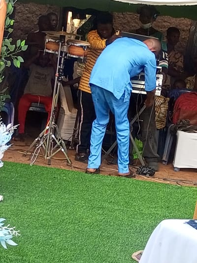 The image depicts an outdoor or semi-enclosed event setup in Amokvo, Nigeria. In the foreground, a vibrant green artificial turf covers the ground, with light blue decorative foliage partially visible on the left and a white tablecloth-draped object on the far right.
The midground features multiple individuals and musical equipment. A man wearing a bright blue traditional Nigerian tunic and trousers, with brown sandals, is centrally positioned, bent forward and facing away from the camera, seemingly adjusting equipment on a black stand. To his left, another man in a yellow and black striped long-sleeved shirt and black trousers is also bent over, interacting with equipment near the first man. Further left, a young man in a light-colored t-shirt and red trousers is seated on a white stool, positioned behind a set of bongo drums mounted on a metallic tripod stand. Cables are visible on the brown ground beneath the equipment.
In the background, several other figures are present, some partially obscured. One individual wears a face mask, and another appears to have headphones on. The background consists of a textured, light brown wall or structure, possibly made of stone or earth. Above, a green canopy or tarp provides overhead shelter.