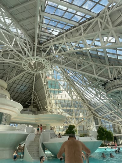 The image depicts the interior of Caldea, a thermal spa in Les Escaldes, Andorra. The foreground shows a waterslide with people on and around it. Some individuals are standing, walking, or sitting on the structure, while others are in the water below. In the background, there is an intricate white steel framework and glass ceiling that allows natural light to enter. The building structure and the sky outside the glass can be seen.