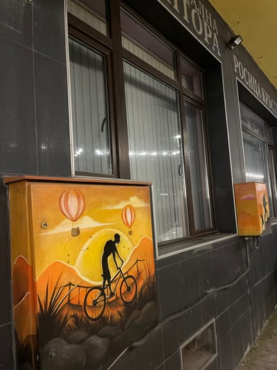 The image shows an outdoor scene in the city of Belogradchik, Bulgaria, focusing on a dark tiled wall with several windows. A large electrical box, painted with a vibrant mural of a cyclist against a sunset with hot air balloons, is prominently featured in the foreground on the left. To the right, a smaller, similarly painted electrical box depicts silhouettes of people against an orange and yellow sky with mountains and clouds. The wall above the windows has text in a light color against a dark background, partially visible. The overall environment appears to be an urban or commercial area, possibly a street or pathway, with a hint of unkempt ground on the right.