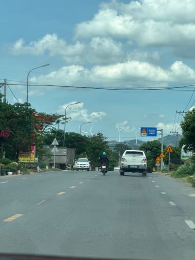 The image depicts a roadside scene in Lao Bảo Commune, Vietnam, viewed from a vehicle. The perspective is tilted, suggesting the photo was taken from a moving car or motorcycle. The road, paved with asphalt, has dashed yellow lines marking the lanes.
Several vehicles are visible: a white pickup truck with the license plate "74C-030.16" is in the foreground, and a motorcycle with a rider wearing a helmet is to the left of the truck. Further ahead, a larger truck is partially visible.
Along the roadside, there are numerous signs. One prominent blue sign indicates "CHÚ Ý GIẢM TỐC ĐỘ QUA KHU VỰC TRƯỜNG HỌC" (Warning: Reduce speed in school zone) with an arrow pointing to the left and "450m". Several yellow triangular warning signs are also present.
The setting is rural or semi-rural, with lush green trees and vegetation lining the road. In the background, hills are visible, and several wind turbines can be seen on the horizon, suggesting the presence of a wind farm. Power lines run overhead, supported by utility poles.
The sky is bright blue with scattered white clouds, indicating a clear, possibly sunny day. The presence of flowering trees with red blossoms adds a touch of color to the scene. Advertisements or business signs are visible at the lower right, one featuring the text "LAT SUAT TAP" and phone numbers. The overall atmosphere is that of a typical day on a Vietnamese road, with a mix of traffic, infrastructure, and natural scenery.