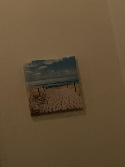 A square canvas print of a beach scene is mounted on a light brown wall. The print depicts a sandy beach with footprints and a few vertical posts. A calm body of water separates the beach from a blue sky with scattered white clouds. The image appears to be taken at an angle, with the bottom left corner of the print closer to the viewer than the top right. The overall lighting is dim and warm, suggesting an indoor setting. The specific location within Englewood, United States, cannot be determined from the image alone.
