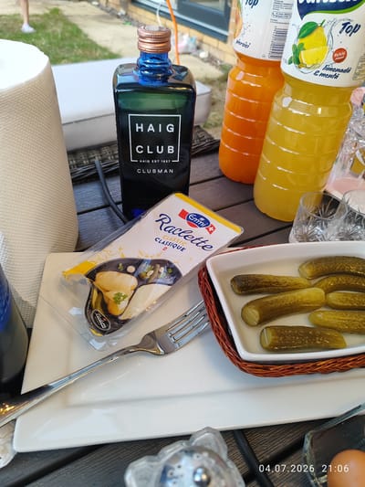 An overhead close-up shot shows an outdoor picnic table setting with various food and drink items. In the center, a dark blue bottle of Haig Club Clubman Scotch Whisky is prominently displayed next to a package of Emmi Raclette cheese. To the right, two plastic bottles of juice, one orange and one yellow (lemonade and mint), stand next to two clear, faceted glasses. In the foreground, a white rectangular dish holds several dill pickles. A fork rests on a white plate beside the dish. In the background, a roll of white paper towels is visible on the left. The scene is set on a dark wooden table, with a wicker basket holding the pickles. The general environment suggests a sunny outdoor gathering. The image was taken at 21:06 on July 4, 2026, in Wellingborough, United Kingdom.