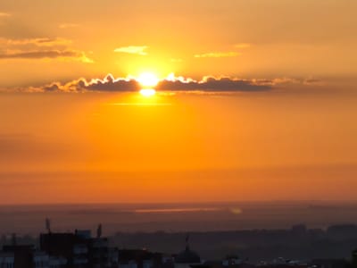 This image captures a vibrant sunset over the city of Svishtov, Bulgaria. The sky is a brilliant orange, transitioning to yellow near the horizon, with streaks of pink and gold. Several clouds are scattered across the sky, some dark and silhouetted against the sun, others illuminated by its rays. The sun itself is a bright, diffused orb, partially obscured by clouds, casting a warm glow across the scene. In the foreground, the dark silhouettes of buildings and structures are visible, suggesting an urban landscape. The horizon line is indistinct, with layers of hazy, muted colors suggesting distant landforms or a body of water. The overall impression is one of a serene and atmospheric evening.